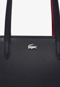 Tote in pelle blu navy con superficie testurizzata, caratterizzato da un logo in coccodrillo argentato e accenti rossi all'interno. Doppie maniglie per il trasporto.