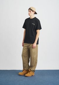 Camiseta negra con un pequeño logo en el pecho, pantalones khaki holgados y botas de trabajo marrones. El fondo presenta una simple pared blanca y un suelo azul.