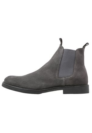 Bottes basses en daim gris avec panneaux élastiques latéraux, languettes de tirage, et une semelle en caoutchouc noire basse. Texture lisse avec des détails de couture contrastants.