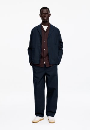 Homme debout face à nous portant une veste et un pantalon bleu marine, une chemise marron à boutons, un sous-vêtement blanc et des baskets blanches à semelles en gomme.