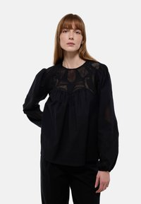Blouse noire avec un empiècement en maille texturée, manches bouffantes longues et devant plissé. Présente des motifs détaillés le long de l'encolure.