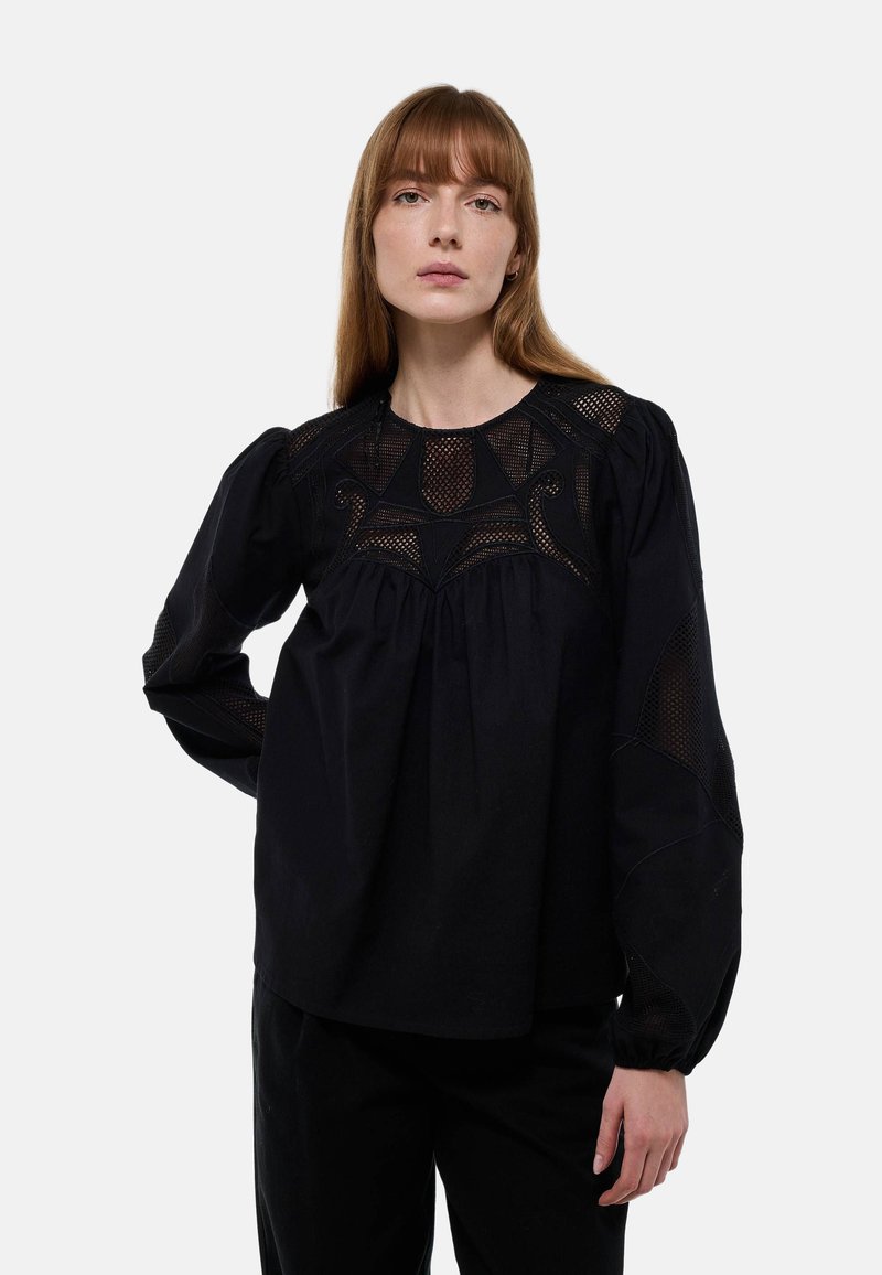 Blouse noire avec un empiècement en maille texturée, manches bouffantes longues et devant plissé. Présente des motifs détaillés le long de l'encolure.