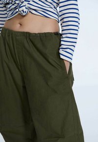 Pantaloni verde oliva con una vestibilità rilassata, dotati di un elastico in vita e tasche laterali, abbinati a una maglietta a maniche lunghe blu e bianca a righe.