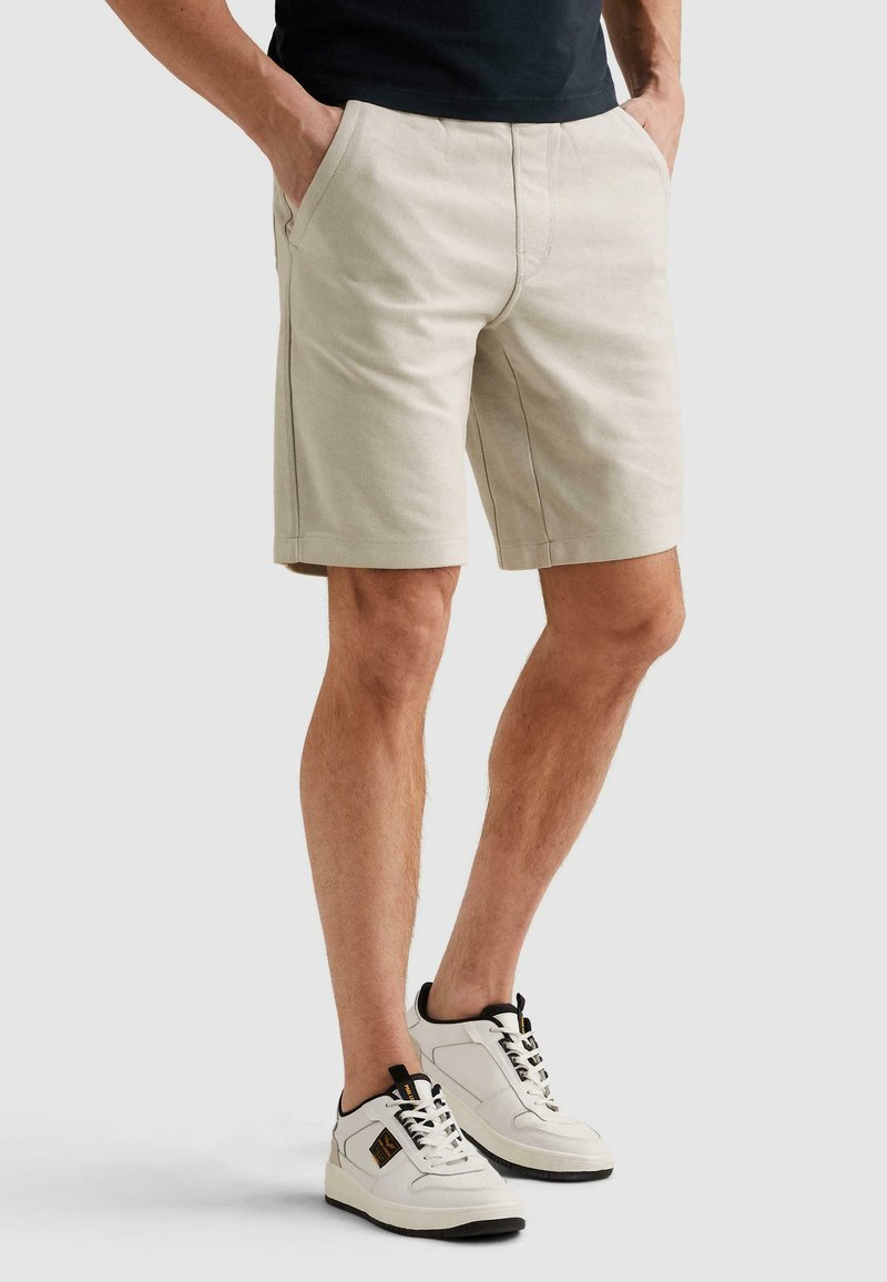 Homme portant un short chino beige et des baskets blanches, debout les mains dans les poches devant un fond uni.