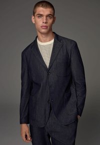 Dunkelblaue Jeans-Blazer mit zwei Vorderseitentaschen, mit Reverskragen und einem Ein-Knopf-Verschluss, getragen über einem gestreiften Hemd. Glatte Textur.