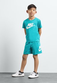 Tyrkysové tričko s krátkým rukávem Nike a odpovídající šortky s bílým logem, doplněné bílými a černými teniskami na šedém povrchu.