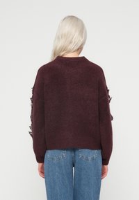 Burgundowy sweter o teksturowym wykończeniu, z luźnym krojem i koronkowymi zdobieniami na rękawach. W połączeniu z jasnoniebieskimi dżinsami.