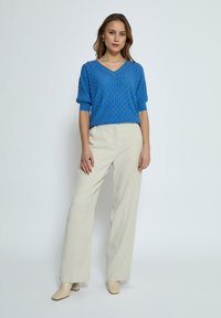 Modrý pletený svetr s výstřihem do V a texturovaným vzorem, spárovaný s krémovými kalhotami s širokými nohavicemi a béžovými lodičkami na blokovém podpatku.