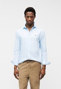 Άνδρας που φορά ανοιχτό μπλε πουκάμισο με κουμπιά και μανίκια που έχουν γυριστεί, και μπεζ παντελόνι κορντόι, στέκεται μπροστά σε απλό λευκό φόντο.