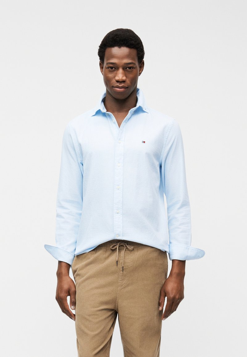 Άνδρας που φορά ανοιχτό μπλε πουκάμισο με κουμπιά και μανίκια που έχουν γυριστεί, και μπεζ παντελόνι κορντόι, στέκεται μπροστά σε απλό λευκό φόντο.