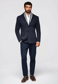 Blazer azul marino con pantalones a juego, camisa blanca y una bufanda estampada. Zapatos de cuero marrón, corte estructurado y dos bolsillos frontales en el blazer.