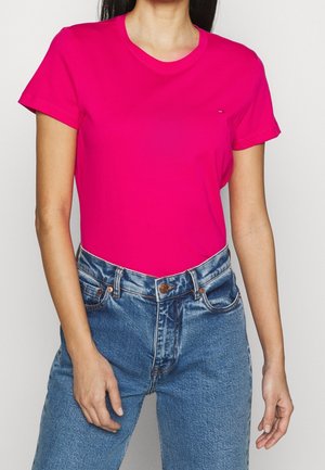 Kvinde iført en lys pink kortærmet T-shirt og blå højttaljede cowboybukser, stående foran en ensfarvet baggrund.