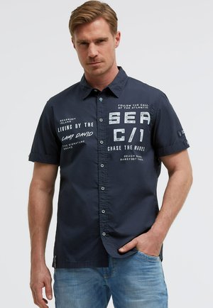 Mann mit kurzen hellbraunen Haaren trägt ein dunkelblaues kurzärmeliges Hemd mit weißem Aufdruck und hellblaue Jeans.