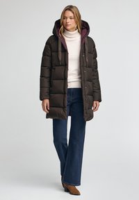 Mörkgrön puffjacka med huva, med en kontrasterande lila foder, tryckknappar och sidofickor, ihop med blå utsvängda jeans.