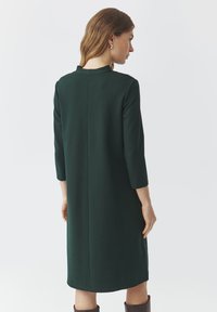 Robe de couleur vert foncé, longueur genoux, fabriquée en tissu lisse. Présente des manches trois-quarts et un design minimaliste avec un détail de couture au dos.