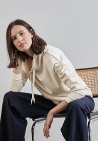 Cream-colored sweatshirt with a cowl neck and drawstring, worn with navy wide-leg pants. Sitting on a chair with rattan accents.