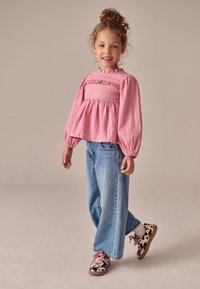 Roze textuurige top met lange mouwen en een gerimpelde taille, gecombineerd met lichtblauwe jeans met wijde pijpen en bruine sneakers met een donker patroon.