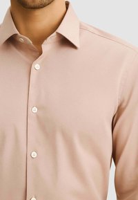 Camicia button-up di colore rosa chiaro, realizzata in tessuto morbido, con colletto a punta e bottoni bianchi in plastica, dal design aderente.