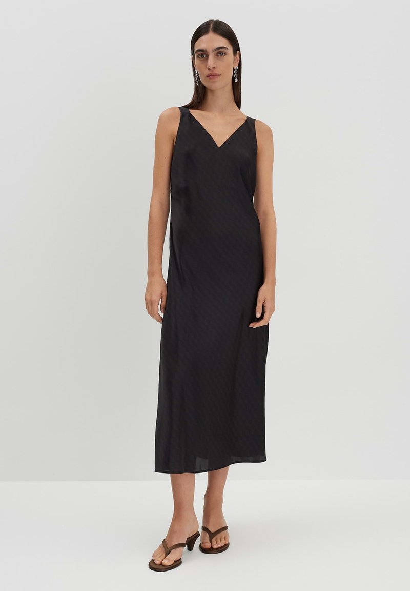 Schwarzes Midikleid mit V-Ausschnitt, aus glattem Stoff, mit einem dezenten Muster. Getragen mit bräunlichen, gestreiften Flip-Flops mit Absatz. Minimalistisches Design.