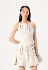 Une robe en lin beige avec des plis, un corsage ajusté et des fines bretelles, s'évasant en une jupe séduisante. Le tissu texturé ajoute un intérêt visuel.