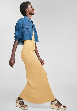 Denim cropped jacket over a ribbed, mustard-yellow maxi dress; accented with gold wedge sandals. Smooth texture and fitted design.