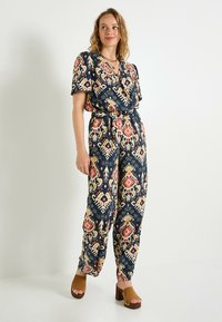 Combinaison marine avec des motifs géométriques colorés, manches courtes, décolleté en V et ceinture à la taille, associée à des sandales à plateforme marron.