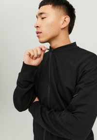 Črna zip-up jakna z visokim ovratnikom in rebrastimi manšetami, ki se ponaša z gladko teksturo in minimalističnim dizajnom, z zadrgo zaprtim žepom.