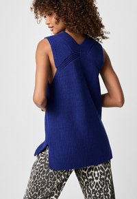 Gebreide mouwloze top in levendig blauw, met een gestructureerd patroon, brede armsgaten en zijsplitten. Gecombineerd met een broek met luipaardmotief.