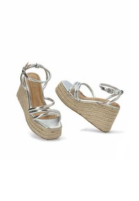 Sandalias de cuña con base de yute trenzado y correas metálicas plateadas. Textura suave, diseño de punta abierta y hebilla ajustable en el tobillo.