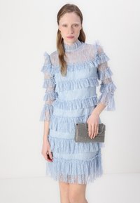 Abito in pizzo azzurro chiaro con volant a strati e maniche trasparenti. Tiene una piccola borsa a tracolla nera e argento, con texture. Sfondo semplice.