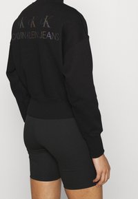 Sudadera negra con puños y dobladillo acanalados, con letras de logo multicolores en la parte trasera. Combinada con pantalones cortos negros.