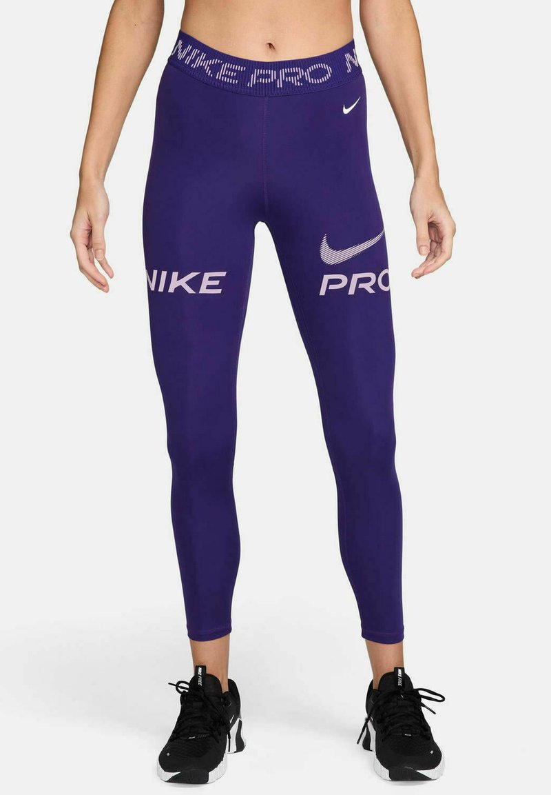 Mujer vestida con leggings Nike Pro morados y zapatillas Nike negras, de pie con los pies a la altura de los hombros contra un fondo blanco.