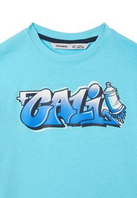 T-shirt azzurro chiaro a maniche corte con scollatura rotonda, caratterizzato da una stampa grafica con la parola "CALI" in diverse tonalità di blu e una bomboletta spray.