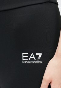 Fekete sportos leggings sima, rugalmas anyagból, fehér EA7 Emporio Armani logóval a combon, testhezálló kivitelben.