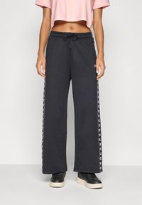 Calças de sweatpants largas e pretas com riscas laterais que apresentam um padrão de logotipo, cintura com cordão e material de algodão suave. Combinadas com sapatilhas pretas.