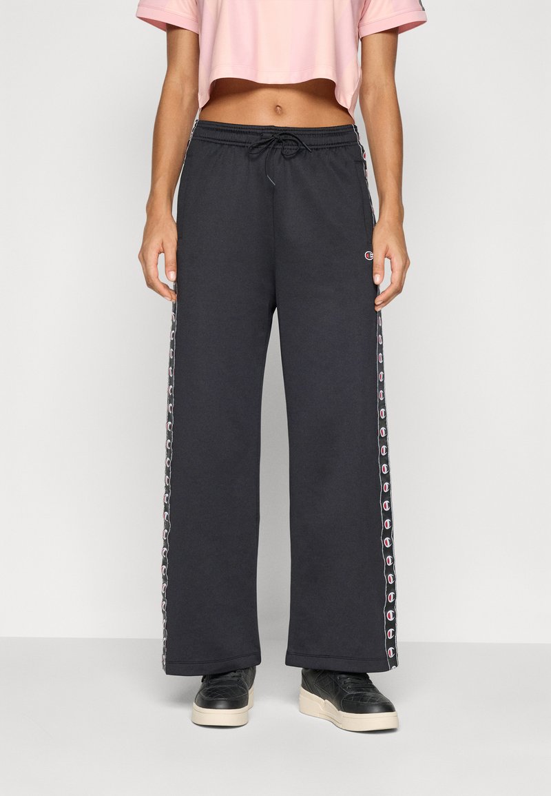 Calças de sweatpants largas e pretas com riscas laterais que apresentam um padrão de logotipo, cintura com cordão e material de algodão suave. Combinadas com sapatilhas pretas.