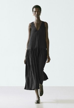 Femme grande marchant vers l'avant, portant une robe maxi sans manches noire texturée, avec des boucles d'oreilles dorées, un bracelet et des chaussures plates foncées, sur un fond uni.