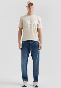 T-shirt grafico beige con testo bianco, jeans in denim blu e sneakers chiare. Modello in piedi su uno sfondo neutro.