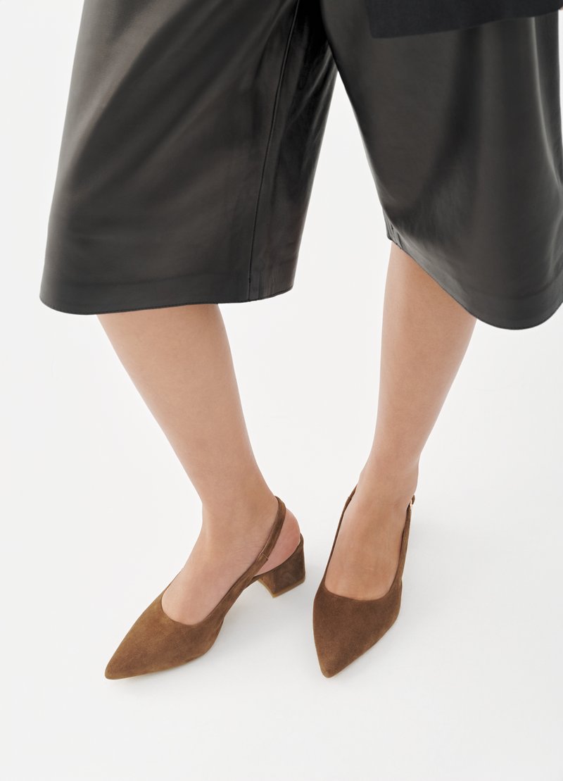 Beine in schwarzen Lederweite-Hosen bis über den Knöchel und braune Wildleder Slingback-Schuhe mit Blockabsatz auf weißem Hintergrund.