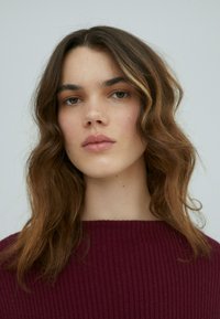 Ein Modell mit welligem, schulterlangem Haar; trägt einen burgunderfarbenen, ribbeligen Pullover. Der Hintergrund ist schlicht und hell, was das Motiv betont.