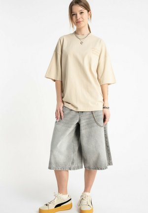 Młoda kobieta w beżowej oversize koszulce, szarych szerokich szortach dżinsowych z łańcuchowym detalem oraz kremowych trampkach z grubymi brązowymi podeszwami.