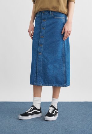Persoon die een blauwe denim midirok met knopen aan de voorkant draagt, witte enkelsokken, zwart-witte lage sneakers en een bruin sweatshirt.