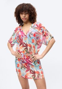 Γυναίκα με σγουρά μαλλιά, φορώντας ένα πολύχρωμο κοντό floral φόρεμα, στέκεται με τα χέρια στη μέση μπροστά σε ένα απλό φωτεινό φόντο.