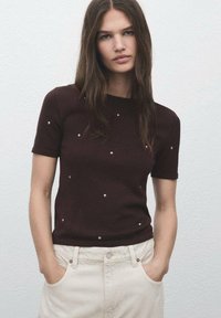 Top in maglia marrone a maniche corte con scollatura rotonda, caratterizzato da borchie argentate sparse; indossato con pantaloni chiari a vita alta.