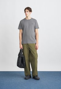 T-shirt cinza de algodão com um pequeno gráfico, calças cargo verde-oliva com bolsos, casaco acolchoado preto e calçado preto. Conjunto sobre um tapete azul.