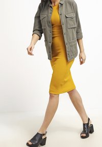 En person bär en senapsgul ribbad midiklänning och en olivgrön oversized skjorta. Skorna är svarta öppna klackar med kraftiga klackar.