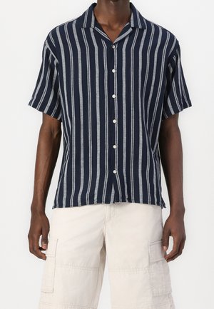 Homme portant une chemise bleu marine à manches courtes avec des rayures blanches verticales et un short cargo beige clair.