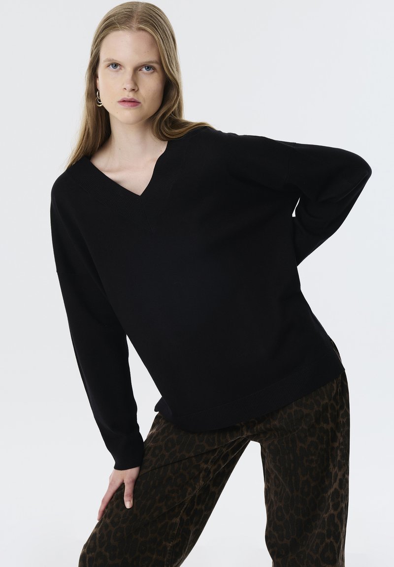 Černý oversized svetr s hlubokým výstřihem do V a žebrovanými manžetami, ve spojení s tmavými kalhotami s leopardím potiskem a jemnou texturou.