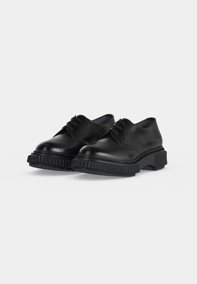 Paire de chaussures en cuir noir à lacets avec des semelles épaisses en caoutchouc rainuré sur un fond clair uni.