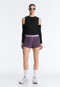 Kobieta ubrana w czarną bluzkę z odkrytymi ramionami na długim rękawie, fioletowe szorty ze ściągaczem, białe skarpetki, szare buty do biegania i okulary przeciwsłoneczne, stojąca na białym tle.